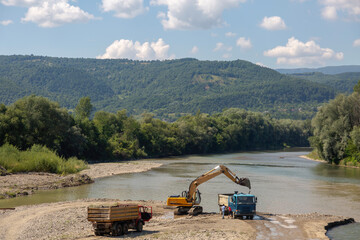 Industrial activity along a river bank: an excavator loads material into trucks, set against a backdrop of rolling forested hills