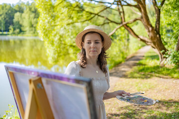 A female artist in a light dress and a straw hat, standing at an easel with a palette in her hands....