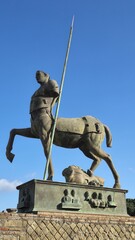 Pompeii, Italy - 8 January 2025. A centaur statue with a raised leg and spear is set on a block featuring busts and faces, viewed against a bright blue sky.