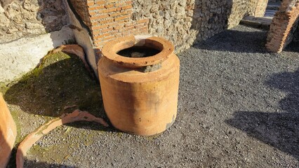 Pompeii, Italy - 8 January 2025. A partially broken terracotta dolium is set in the floor beside ancient masonry, likely part of a Roman shop or kitchen.