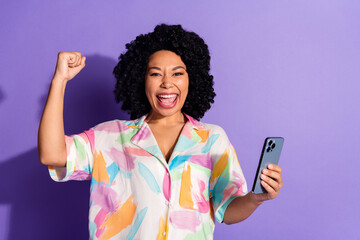 Cheerful young woman holding a smartphone celebrating success on a vibrant purple background in a...