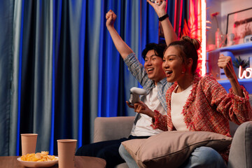 Couple gamer raising fist up holding joysticks be winner together complete level sitting sofa playing video game with front snack and drinks at neon light color living room at modern home. Infobahn.