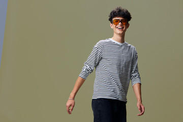 Smiling young man wearing a striped shirt and orange sunglasses, standing in a relaxed pose with...
