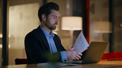 Obraz premium Late worker reading papers working at laptop in dark office closeup. Serious man