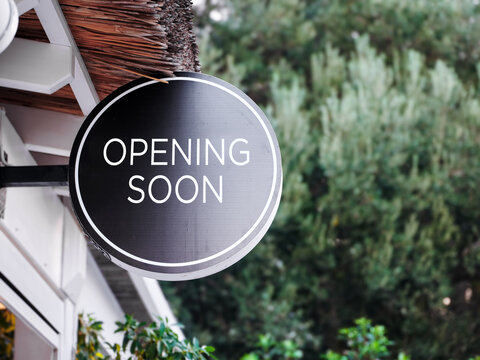 Round black sign on a shop with text opening soon is hanging on a building