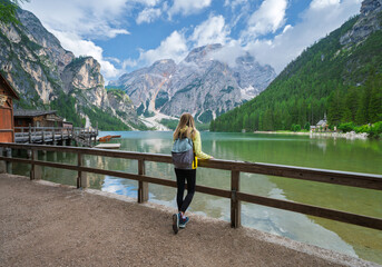 Young woman with backpack on the Braies lake in alpine mountains, blue sky with clouds, reflection in clear water, wooden fence at sunset. Summer travel in Dolomites, Italy. Girl on the lake. Hiking