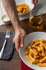 Vegan pasta with dried tomato served on a white deep plate by a hand, viewed from a 45 degree angle. Next to the dish sits a fork with a grey tissue, a glass of white wine, a small bolw with parmesano