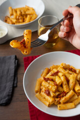 hand picking vegan pasta with dried tomato served on a white deep plate, viewed from a 45 degree angle. Next to the dish sits a glass of white wine, a small bowl with parmesano and the hint of another