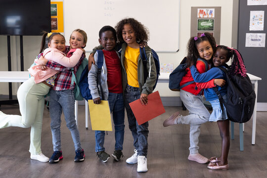 Happy diverse schoolchildren hugging and smiling in classroom, holding colorful folders