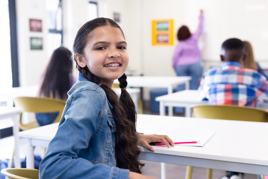 Smiling girl in classroom looking back, holding pencil, female teacher writing on board, at school