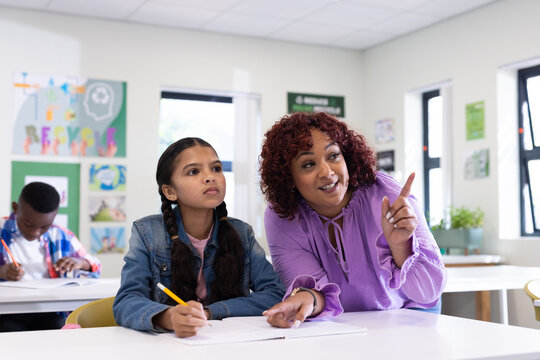 Female teacher guiding girl with pencil in classroom, focusing on learning together, at school
