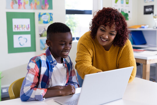 Female teacher helping african american boy with laptop in classroom, both smiling, at school