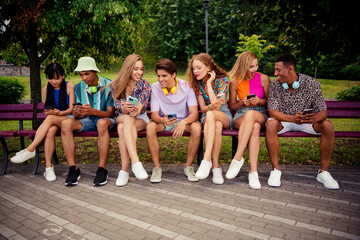 Buddies sitting together on a bench outdoors exploring smartphones and enjoying summer day