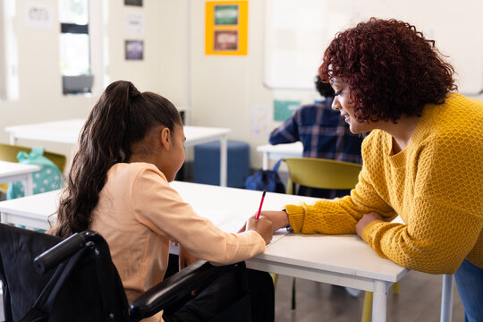 Female teacher assisting girl in wheelchair with writing task in classroom, at school