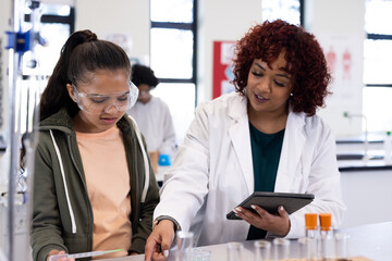 Female teacher guiding girl in science lab, using tablet for experiment instructions, at school