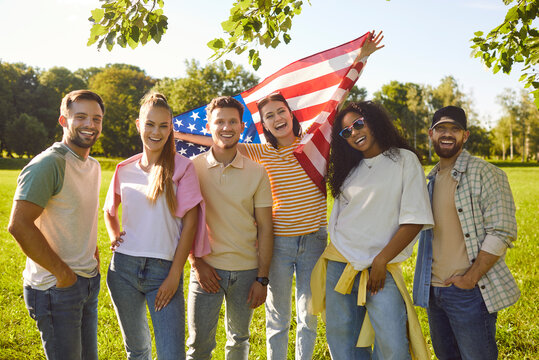 Portrait of happy smiling young people friends in casual clothes holding USA flag standing together in summer park and looking cheerful at camera outdoors. Independence Day celebration concept.