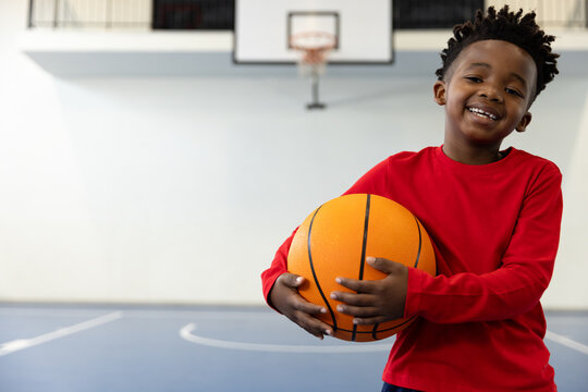 Smiling african american boy holding basketball in school gym, enjoying sports activity, copy space - Powered by Adobe