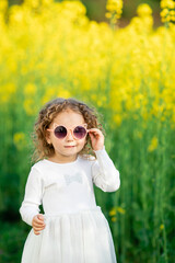  A little girl in a white dress and cat-shaped pink sunglasses stands by a bright yellow flower...