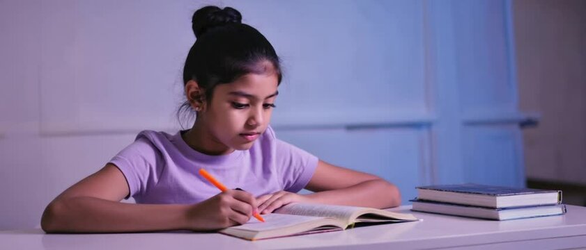A diligent Latina girl writes in textbook under blue evening light beside stacked volumes. Concept of determined late-night learning.
