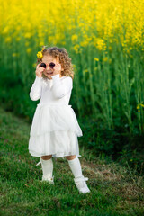 A little girl in a white dress and cat-shaped pink sunglasses stands by a bright yellow flower...