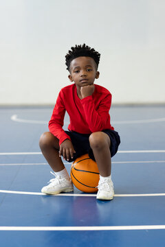 Young african american boy sitting on basketball in gym, wearing red shirt, looking thoughtful