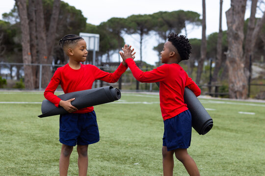 Diverse boys high-fiving outdoors holding yoga mats, enjoying teamwork and exercise - Powered by Adobe