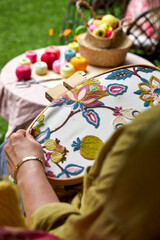 Over-the-shoulder close-up view of a woman embroidering a pattern on fabric held in an embroidery hoop. The hands and hoop are in focus, while her shoulder and back are softly blurred in the foregroun