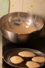 A bowl of pancake batter next to a pan with pancakes cooking on a stove, illustrating the process of making pancakes in a kitchen setting