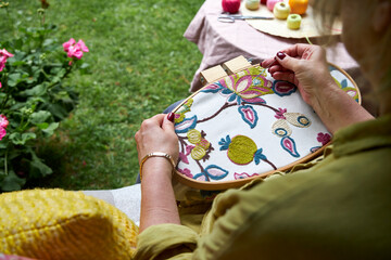 Front close-up view of a woman's hands embroidering a decorative pattern on fabric stretched in an embroidery hoop. The hands are focused and steady, with visible stitches forming on the fabric. Natur