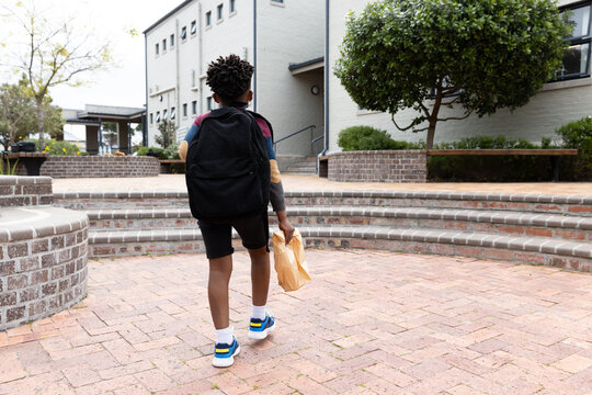 African american boy with backpack walking towards school building, holding lunch bag