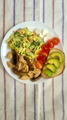 Healthy balanced meal on a plate featuring bread with avocado, tomatoes, mozzarella, cabbage and carrot salad, chicken pieces and mushrooms placed on a tablecloth with stripes .