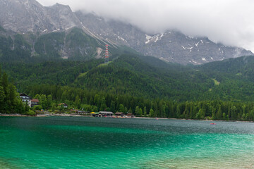 A serene alpine lake with crystal-clear turquoise water mirrors the snow-capped Zugspitze and pine-covered hills. Rowboats glide gently under a clear blue sky—pure alpine tranquility.