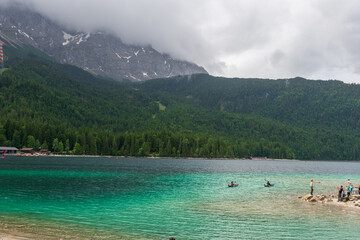 A serene alpine lake with crystal-clear turquoise water mirrors the snow-capped Zugspitze and pine-covered hills. Rowboats glide gently under a clear blue sky—pure alpine tranquility.