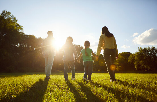 Family fun walking together in grassy green field, close holding hands, moving towards sun shining brightly spending time together outdoors, enjoying loving kind moment and scenery, rear view