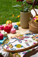 Close-up of a small garden table with embroidery supplies on a warm summer day. Colorful threads, needles, fabric, scissors, and a wooden hoop are neatly arranged in natural light. The table stands in