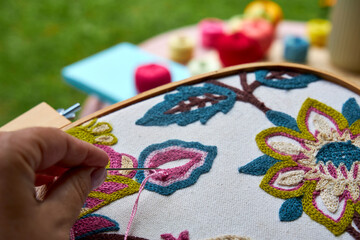 Front close-up view of a woman's hands embroidering a decorative pattern on fabric stretched in an embroidery hoop. The hands are focused and steady, with visible stitches forming on the fabric. Natur