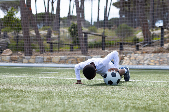 Young african american boy practicing push-ups on soccer field with ball, building strength