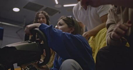 Friends gather around as a young woman plays a racing video game with a steering wheel, reacting to the action on screen.