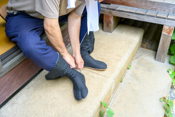 作業着姿の男性が玄関先で地下足袋を履こうと前かがみになって準備を整える真夏の仕事前の一幕
