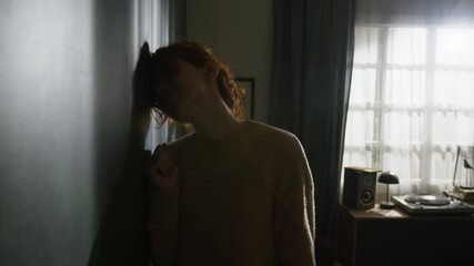 Upset red-haired young woman banging her fists on wall and sliding down onto floor in dimly lit room - Powered by Adobe