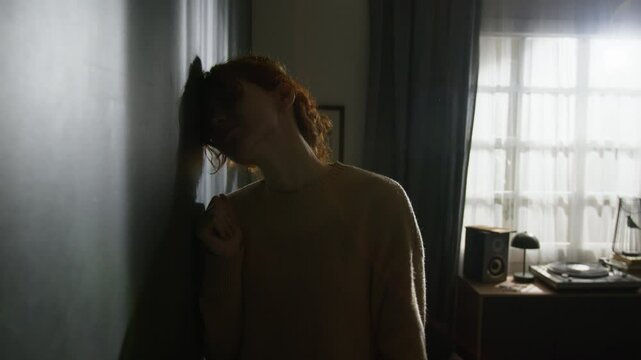 Upset red-haired young woman banging her fists on wall and sliding down onto floor in dimly lit room