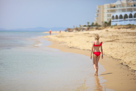 Back of 10-years-old caucasian girl with blonde hair walks along sandy beach in Tunisia. Calm sea and hotel buildings on background. Travel with children concept.
