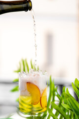 A cold peach-infused drink being poured into a glass filled with ice and fruit slices, surrounded...