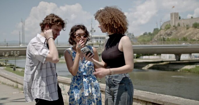 Three friends explore a city, using a smart phone for directions. They discuss their route and point towards landmarks.