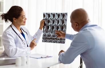 Smiling African American female doctor in medical coat explaining spine x-ray results to attentive...