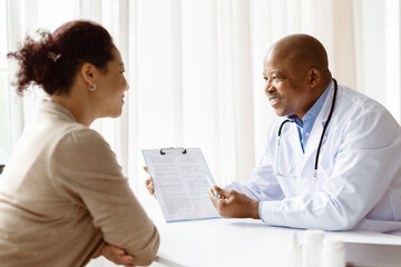Fototapeta premium African American male doctor cheerfully explaining medical documents to black woman patient during positive consultation, professional therapist showing test results to young female in clinic