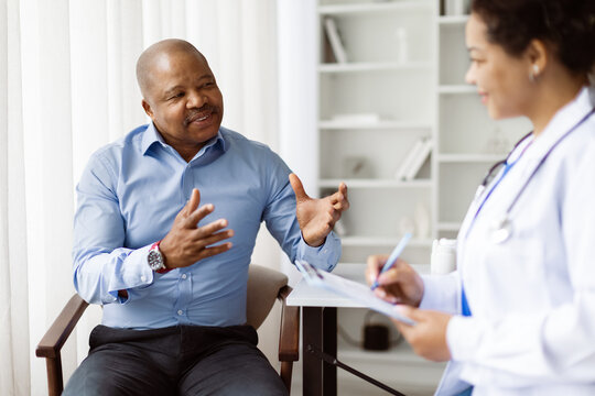 Black Male Patient Talking To Doctor Woman During Meeting In Clinic, African American Mature Man Explaining Health Issues To Therapist Female That Making Records To Clipboard