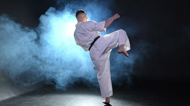 Strong man fighter in white kimono for karate martial arts with black belt preparing for fight training in black background. Man training moves punches kicks practicing shadow fight ready for sparring