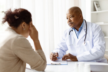 Focused African American male doctor listening carefully while black millennial patient discusses her symptoms, seated in a clinical environment, female explaining health problems to therapist man