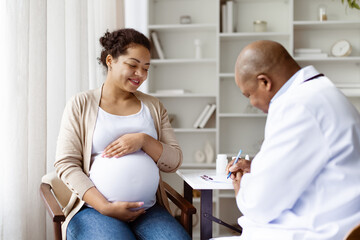 Pregnancy Check Up. Professional black senior obstetrician listening to patient and writing record, happy pregnant lady hugging belly sitting at clinic, having consultation with gynecologist